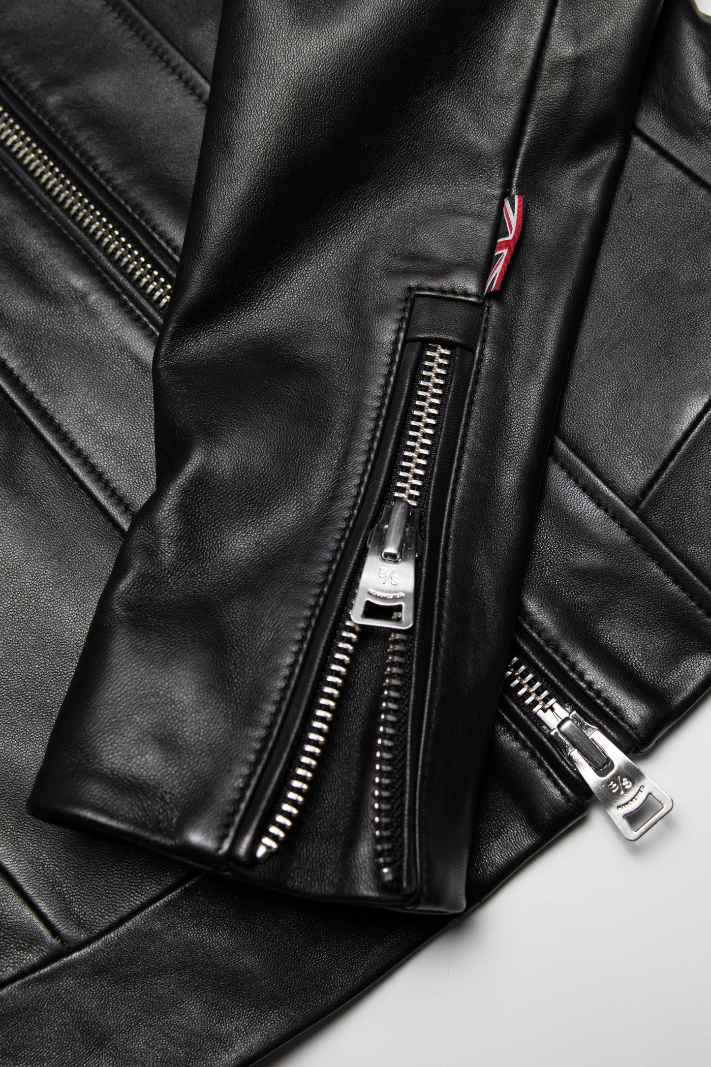 Close up of a Womens black Moto Racer Leather Jacket on a light beige background showing silver hardware detail and a british design flag
