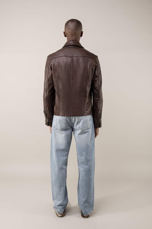 Back shot of a person wearing a brown leather jacket and light blue jeans on a plain background