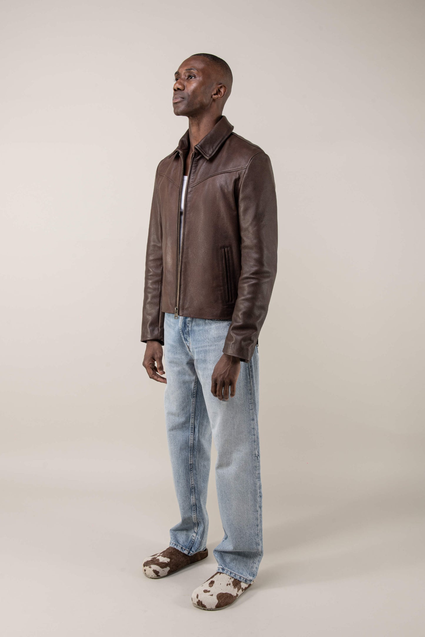 Man wearing a brown leather jacket and light blue jeans on a beige background